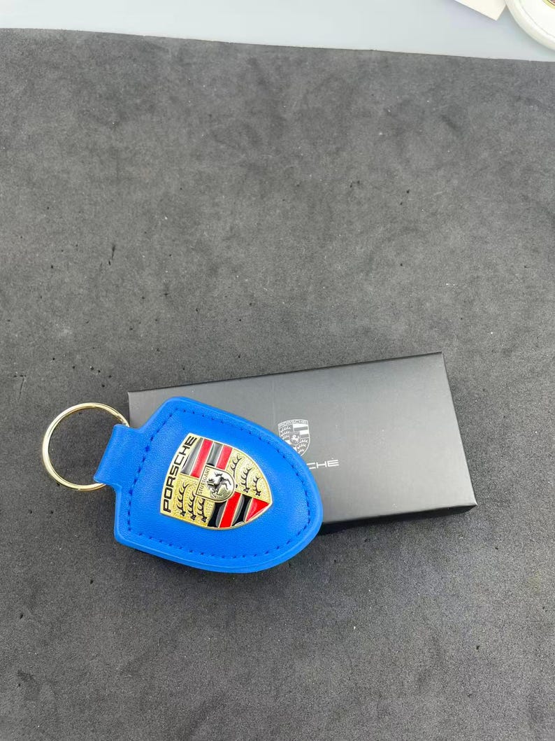 Op de afbeelding: Een felblauwe leren sleutelhanger met een goudkleurig metalen Porsche-embleem met rode en zwarte details. De sleutelhanger is bevestigd aan een ronde goudkleurige metalen ring en ligt naast een kleine zwarte rechthoekige doos.