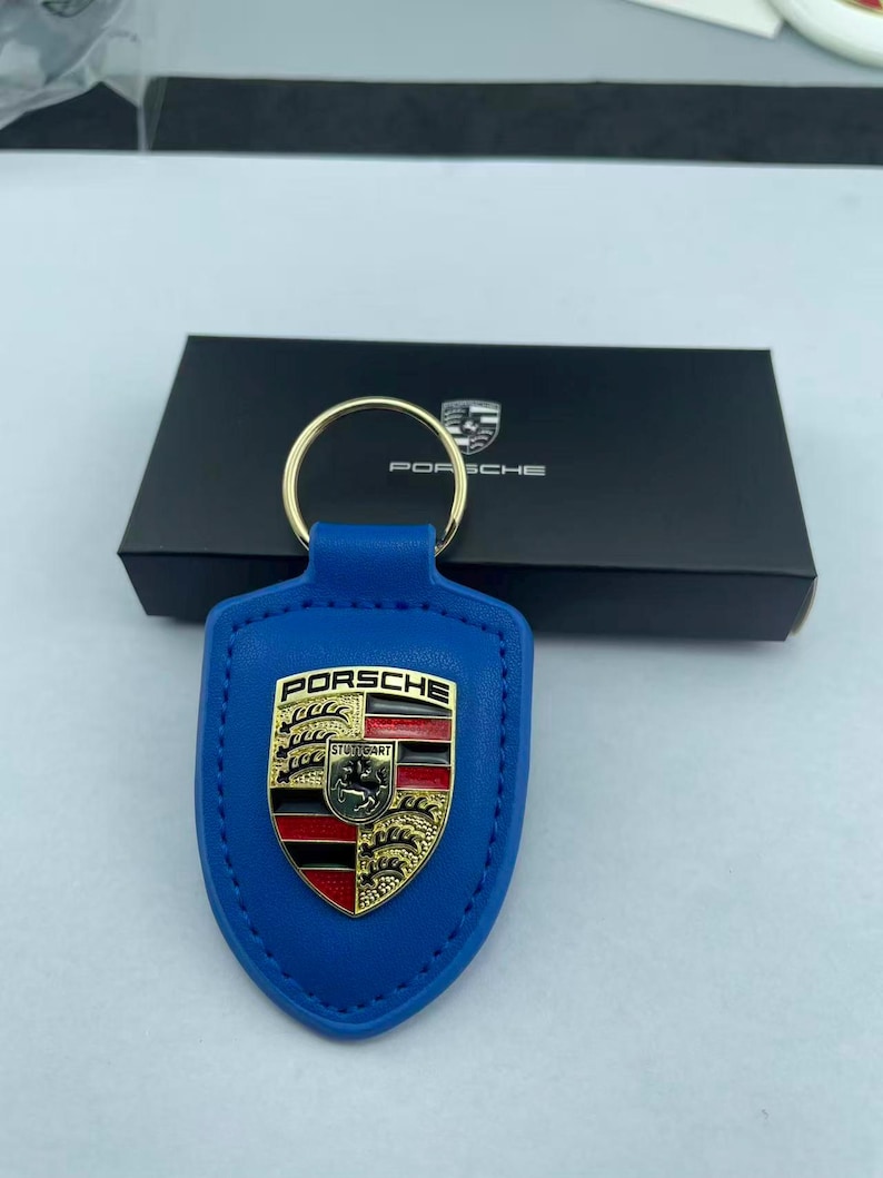 Op de afbeelding: Een blauwe leren sleutelhanger met een goudkleurig metalen Porsche-embleem met rode en zwarte details. De schildvormige hanger is bevestigd aan een goudkleurige metalen ring. Achter de hanger staat een zwarte doos met het Porsche-logo.