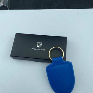 Op de afbeelding: Een felblauwe, schildvormige lederen sleutelhanger met een goudkleurige metalen ring. De sleutelhanger ligt voor een rechthoekige zwarte doos met het witte Porsche-logo en de tekst in het midden van het deksel gedrukt.