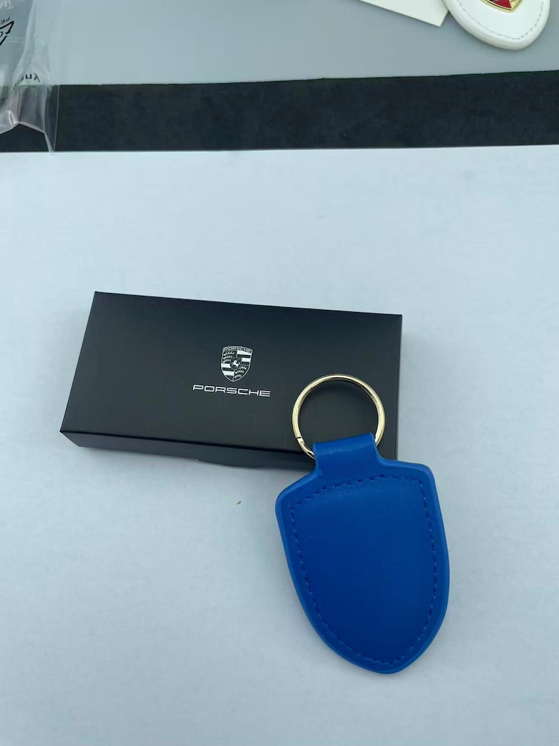 Op de afbeelding: Een felblauwe, schildvormige lederen sleutelhanger met een goudkleurige metalen ring. De sleutelhanger ligt voor een rechthoekige zwarte doos met het witte Porsche-logo en de tekst in het midden van het deksel gedrukt.