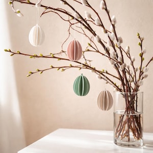 Puede incluir: Una decoración de Pascua. Varios adornos esféricos de colores pastel cuelgan de ramas con amentos blancos. Las ramas están en un jarrón de cristal transparente sobre una superficie blanca. Los adornos son blancos, rosas, verdes y beiges.