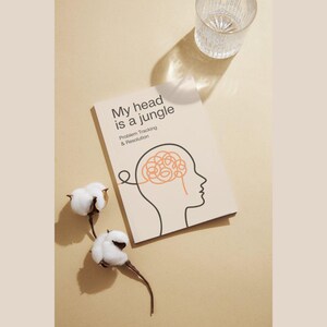 Op de afbeelding: Een boek getiteld "My head is a jungle" met een hersenillustratie, een glas water en katoenstelen op een beige oppervlak. De kaft van het boek toont een lijntekening van een hoofd met een wirwar van hersenen.