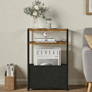 May include: A three-tiered storage shelf with a black metal frame and wooden shelves. The top shelf holds a vase of flowers and a travel mug. The middle shelf has books and a decorative figurine. The bottom shelf has a black fabric magazine holder with a magazine that says "Cereal".