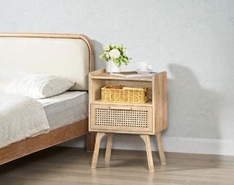 May include: A light wood nightstand with a woven rattan drawer and shelf. A small woven basket, a vase of flowers, and a cup and saucer sit on top. The nightstand is next to a bed with a white comforter and pillow.