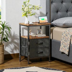 May include: A two-drawer nightstand with a metal frame and a wood top. The drawers are dark gray with black handles. A tablet, a photo frame, and a vase with flowers are on top of the nightstand. A bed with a gray headboard is in the background.