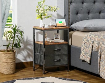 May include: A two-drawer nightstand with a metal frame and a wood top. The drawers are dark gray with black handles. A tablet, a photo frame, and a vase with flowers are on top of the nightstand. A bed with a gray headboard is in the background.