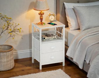 May include: White nightstand with two drawers and a shelf, featuring a lamp, alarm clock, and a phone. The lamp has a beige lampshade and a decorative base. The nightstand is next to a bed with white bedding.