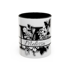 Puede incluir: Taza de cerámica blanca con interior negro. La taza presenta una gran letra "M" negra con detalles florales y mariposas. El nombre "Melissa" está escrito en cursiva en el diseño.