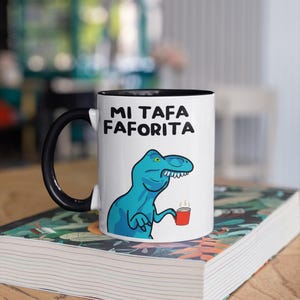 Puede incluir: Taza de cerámica blanca con asa y borde negros, con una ilustración de un dinosaurio azul sosteniendo una taza de café roja. La taza tiene el texto "MI TAFA FAVORITA" en negro. La taza está sobre un libro.