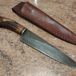 May include: A fixed-blade knife featuring a textured brown antler handle with a metallic bolster and a pointed steel blade, accompanied by a smooth brown leather sheath with visible stitching along the edge of the tapered protective cover.