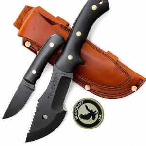 Könnte beinhalten: Ein schwarzes Tracker-Messer mit gezahnter Klinge und ein kleineres passendes schwarzes Messer, beide mit Messingnieten an den Griffen. Eine hellbraune Lederscheide mit Kupfernieten und ein olivgrüner Aufnäher mit Adler-Logo sind enthalten.