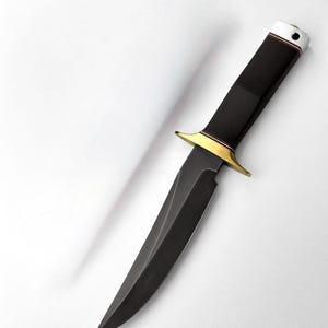 Puede incluir: Un cuchillo de hoja fija con una hoja negra mate y un mango negro a juego. El cuchillo incluye una guarda de metal dorado pulido y un pomo plateado en el extremo del mango, que presenta un pequeño orificio circular para un cordón.
