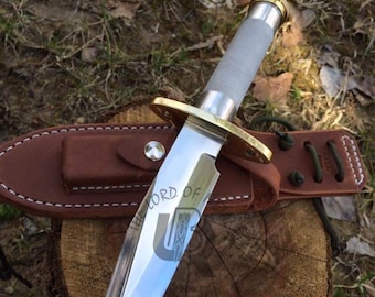 May include: A large, silver-bladed knife with a gray handle and gold accents, resting in a brown leather sheath. The blade has the text "LORD OF" and a logo etched on it. The knife is on a wooden surface.
