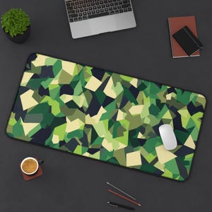 May include: A rectangular desk mat with a geometric camouflage pattern in shades of green, tan, and navy. A white computer mouse rests on the mat. Other items on the dark surface include a laptop, a small potted plant, and stationery.