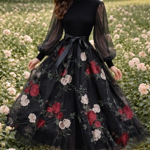 Op de afbeelding: Zwarte jurk met een getailleerd lijfje en doorschijnende lange mouwen. De rok is van tule en versierd met rode en witte bloemenborduursels. Een zwarte satijnen strikriem accentueert de taille. Geschikt voor formele evenementen.