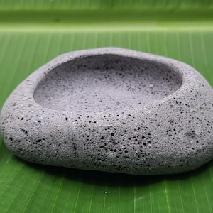 Peut inclure: Un bol en pierre grise et poreuse de forme arrondie. L'intérieur du bol est lisse et l'extérieur texturé. Le bol est posé sur une feuille verte.