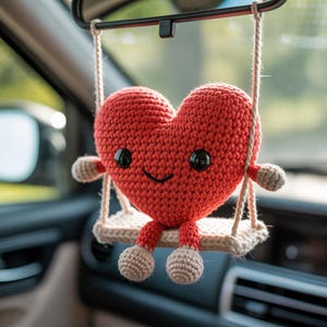 Pode incluir: Um enfeite em forma de cora&ccedil;&atilde;o vermelho de croch&ecirc; com olhos pretos e boca sorrindo, sentado em um pequeno balan&ccedil;o. O balan&ccedil;o &eacute; suspenso por cordas bege, e o enfeite tem pequenos bra&ccedil;os e p&eacute;s. O fundo est&aacute; desfocado.