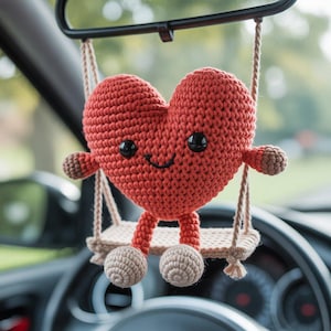 Pode incluir: Enfeite em forma de cora&ccedil;&atilde;o vermelho de croch&ecirc;, com olhos de bot&atilde;o pretos e uma boca sorridente, pendurado em um pequeno balan&ccedil;o. O balan&ccedil;o &eacute; suspenso por cordas bege, e o cora&ccedil;&atilde;o tem pequenos bra&ccedil;os e p&eacute;s. O enfeite est&aacute; pendurado no espelho retrovisor de um carro.