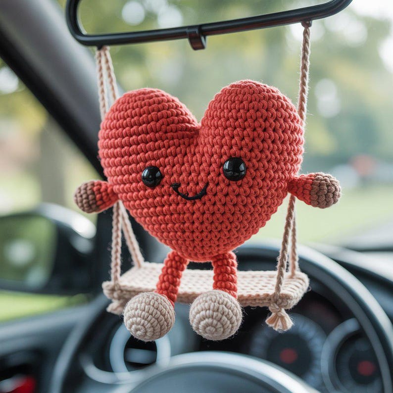 Pode incluir: Enfeite em forma de cora&ccedil;&atilde;o vermelho de croch&ecirc;, com olhos de bot&atilde;o pretos e uma boca sorridente, pendurado em um pequeno balan&ccedil;o. O balan&ccedil;o &eacute; suspenso por cordas bege, e o cora&ccedil;&atilde;o tem pequenos bra&ccedil;os e p&eacute;s. O enfeite est&aacute; pendurado no espelho retrovisor de um carro.