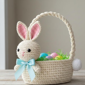 May include: A crocheted Easter basket with a bunny attached. The bunny has pink-lined ears, black eyes, and a light blue bow. The basket contains colourful Easter eggs and green decorative grass. A white pom-pom tail is attached to the basket.