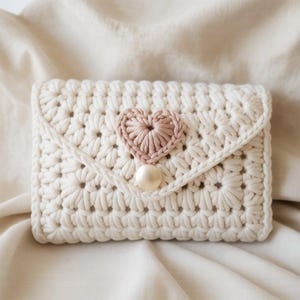 May include: Cream-colored crochet wallet with a flap closure and a light pink heart accent. The wallet is secured with a pearl button. The crochet pattern creates a textured, floral design. The wallet is a handmade item.