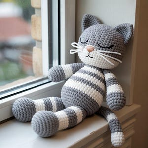 Peut inclure: Poupée chat au crochet faite à la main, rayée grise et blanche, avec les yeux fermés, des moustaches blanches et un nez rose. Le chat est assis sur un rebord de fenêtre blanc. Le corps du chat est gris avec des rayures blanches.