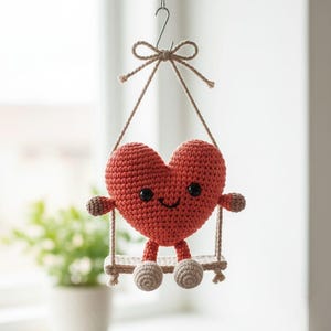 Pode incluir: Um brinquedo de pel&uacute;cia em forma de cora&ccedil;&atilde;o, feito &agrave; m&atilde;o, de croch&ecirc;, com um rosto sorridente, bra&ccedil;os e pernas, sentado em um pequeno balan&ccedil;o. O cora&ccedil;&atilde;o &eacute; de uma cor vermelho-alaranjada vibrante, com detalhes marrons nos bra&ccedil;os e pernas. O balan&ccedil;o &eacute; suspenso por uma corda bege.