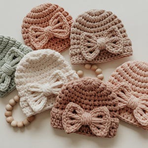 Op de afbeelding: Een verzameling handgemaakte gehaakte baby mutsen in pastelkleuren, waaronder roze, beige en mintgroen. Elke muts heeft een grote, decoratieve strik aan de voorkant. Een houten bijtring is ook zichtbaar.