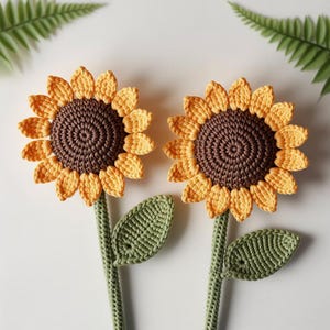 May include: Two handmade crochet sunflowers. The flower heads are yellow and brown, with green stems and leaves. The sunflowers are set against a white background, with a fern leaf in the upper corners.