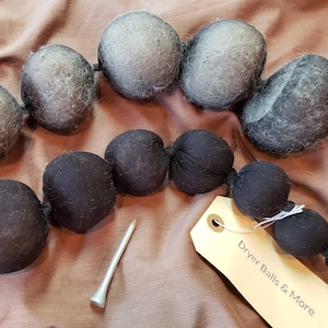Könnte beinhalten: Eine Sammlung grauer und schwarzer Trocknerbälle, einige in Schnüren verbunden, mit einem Golf-Tee und einem Etikett mit der Aufschrift "Dryer Balls & More". Die Trocknerbälle bestehen aus einem faserigen Material.