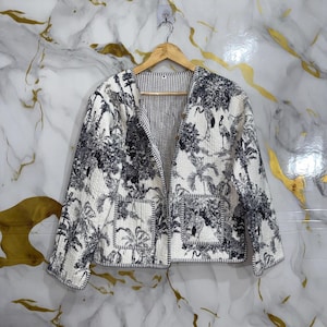 Könnte beinhalten: Eine gesteppte Jacke mit offener Vorderseite und schwarz-weißem Toile-de-Jouy-Druck mit tropischen Palmen und Kranichen. Das Kleidungsstück hat lange Ärmel, zwei aufgesetzte Vordertaschen und einen gestreiften Saum, hängend an einem Holzkleiderbügel.