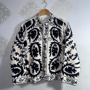 May include: Cream-coloured jacket with intricate black floral embroidery. The jacket features a mandarin collar and a button-up front. The embroidery includes stylized leaves and swirling patterns, creating a detailed and ornate design. The jacket is hanging on a wooden hanger.