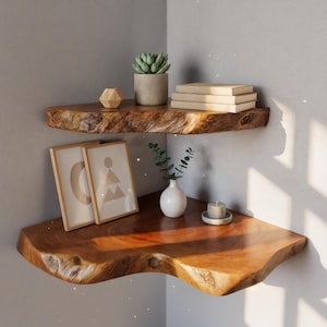 Puede incluir: Dos estanterías de esquina de madera en bruto con varios objetos decorativos. La estantería superior contiene una suculenta en una maceta de hormigón, libros y una escultura geométrica de madera. La estantería inferior muestra arte enmarcados, un pequeño jarrón y una vela.