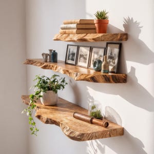 Puede incluir: Tres estanterías flotantes de madera con bordes naturales. Exhiben libros, fotos enmarcadas, plantas en macetas y jarrones decorativos. Montadas en una pared blanca, crean una estética rústica.