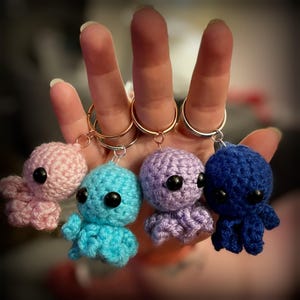 Può includere: Quattro portachiavi a forma di polpo all'uncinetto in rosa, blu, viola e blu navy. Ogni polpo ha grandi occhi neri e un portachiavi color oro. I tentacoli sono visibili e il lavoro all'uncinetto è dettagliato.