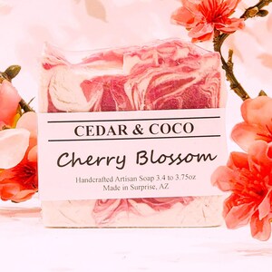 May include: A rectangular bar of handcrafted artisan soap with a pink and white marbled pattern. A white paper label features the text CEDAR & COCO, Cherry Blossom, and Handcrafted Artisan Soap 3.4 to 3.75oz, Made in Surprise, AZ.