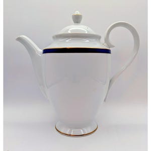 May include: White porcelain teapot with a curved handle and spout. The teapot features a blue band with gold trim around the top and base, and a gold-trimmed lid with a small finial. The design is simple and elegant.