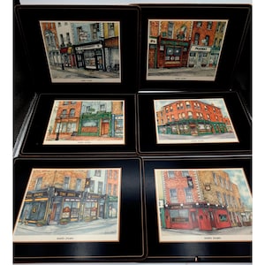 May include: A set of six rectangular placemats with black borders, each featuring a distinct watercolor-style illustration of a storefront or pub. The scenes depict various brick buildings, including establishments labeled as The Duke and James Toner.
