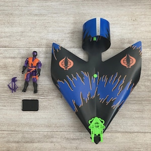 Puede incluir: Un juego de juguetes vintage con un avión negro y azul con detalles en naranja y un accesorio verde. Incluye una figura de acción morada y naranja, un arma pequeña morada y una base rectangular negra. El avión tiene dos logotipos de Cobra naranjas.