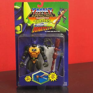 Pode incluir: Figura de ação Donatello da série TMNT Supermutants, embalada em blister azul e verde. A figura é roxa, laranja e verde, com acessórios. A embalagem inclui o texto "TMNT Supermutants" e "Donatello".