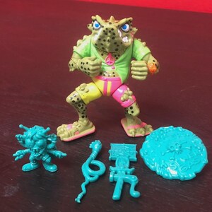 May include: A vintage action figure of a frog-like character in a green shirt, yellow and pink shorts, and sandals. Accessories include a small blue monster, a snake, a sign, and a round blue object.