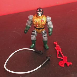 Könnte beinhalten: Vintage Actionfigur einer roboterartigen Figur mit goldfarbenem und grauem Torso, schwarzem Helm und grünen Stiefeln. Zubehör sind ein weißes Kabel und eine rote Waffe. Die Figur steht auf einer dunklen Oberfläche.