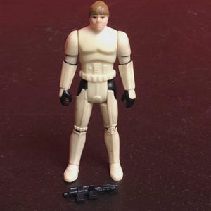 Op de afbeelding: Vintage actiefiguur van een personage in een wit en zwart uniform, met een zwart wapen aan zijn voeten. De figuur heeft een bruin hoofd en bruin haar. De figuur staat op een donkere ondergrond.