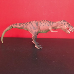 May include: A plastic Tyrannosaurus Rex toy in shades of brown and green, with dark spots and a long tail. The dinosaur has a detailed head with visible teeth and is posed on a black surface against a red background.