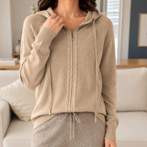 May include: A beige knit zip-up hoodie featuring a vertical cable knit pattern along the front zipper closure, long sleeves with ribbed cuffs, a matching drawstring hood, and a ribbed hemline, crafted from a soft, textured knit fabric.