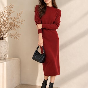 May include: A long-sleeved, calf-length sweater dress in a deep red hue, featuring a high neckline and ribbed cuffs. The garment is paired with a small black handbag and black ankle boots, creating a cohesive look with simple lines.