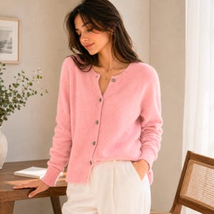 May include: A soft, light pink long-sleeved cardigan featuring a round neckline and a vertical row of small, dark buttons down the front. The knit garment has ribbed cuffs and a ribbed hem, providing a textured appearance with a classic button-down design.