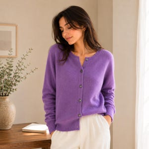 May include: A vibrant purple long-sleeve cardigan featuring a round neckline and a front button-down closure with matching purple buttons. The knit garment has ribbed cuffs and a ribbed hem, providing a textured finish to the soft, solid-coloured fabric.