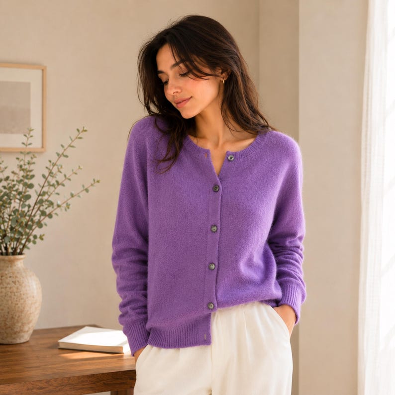 May include: A vibrant purple long-sleeve cardigan featuring a round neckline and a front button-down closure with matching purple buttons. The knit garment has ribbed cuffs and a ribbed hem, providing a textured finish to the soft, solid-coloured fabric.