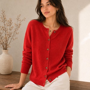 May include: A vibrant red long-sleeved knit cardigan featuring a round neckline and a vertical row of small, round, dark-coloured buttons down the front. The garment has ribbed cuffs and a ribbed hem, providing a structured fit with a soft texture.